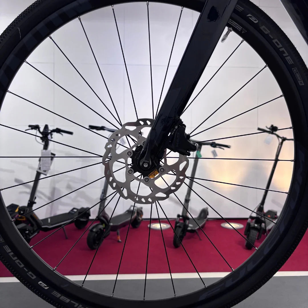 Izdelek na sliki je električno kolo, znamke Raymon, model GravelRay E 7.0. Na sliki je prikazan sprednji del kolesa, kjer so vidni platišče, žice in disk zavore, v ozadju pa so vidni električni skiroji. Kolo je zasnovano za vožnjo po različnih terenih.