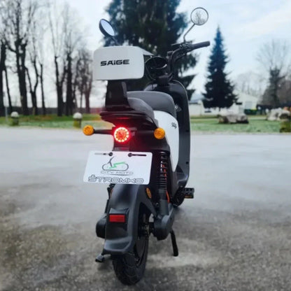 Na sliki je električni skuter znamke Saige, model Stromko. Vidna je zadnja stran skuterja, ki ima elegantno obliko z LED lučmi in registracijsko tablico. Okolica je mirna, z drevesi v ozadju, kar daje občutek prostornosti in narave.