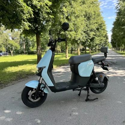 Na sliki je električni skuter, znamke Stromko. Ima svetlo modro barvo in sodoben dizajn, s širokim sedežem in udobnim položajem za vožnjo. Skuter je parkiran na poti, obdan z drevesi, kar nakazuje na prijetno okolje za vožnjo. Vidni so tudi zadnji ogledali in sprednji del skuterja, kar poudarja njegovo funkcionalnost.