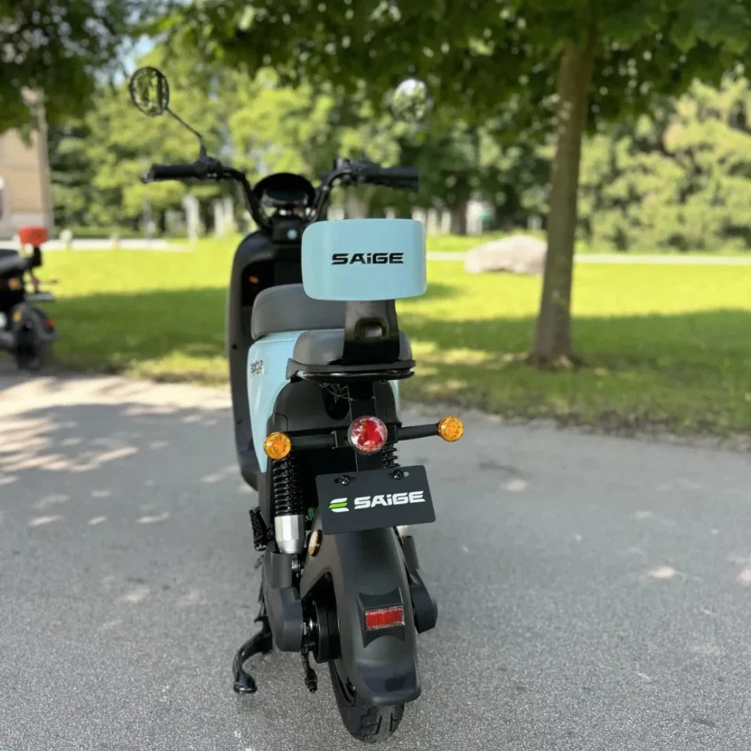 Na sliki je električni skuter znamke SÄIGE. Vidna je zadnja stran skuterja, ki ima modro-sivo barvno kombinacijo in je opremljen z zadnjimi lučmi ter registracijsko tablico. Okolica je zelena, kar daje občutek prostornosti in narave. Skuter je zasnovan za mestno vožnjo in je praktičen za vsakodnevno uporabo.