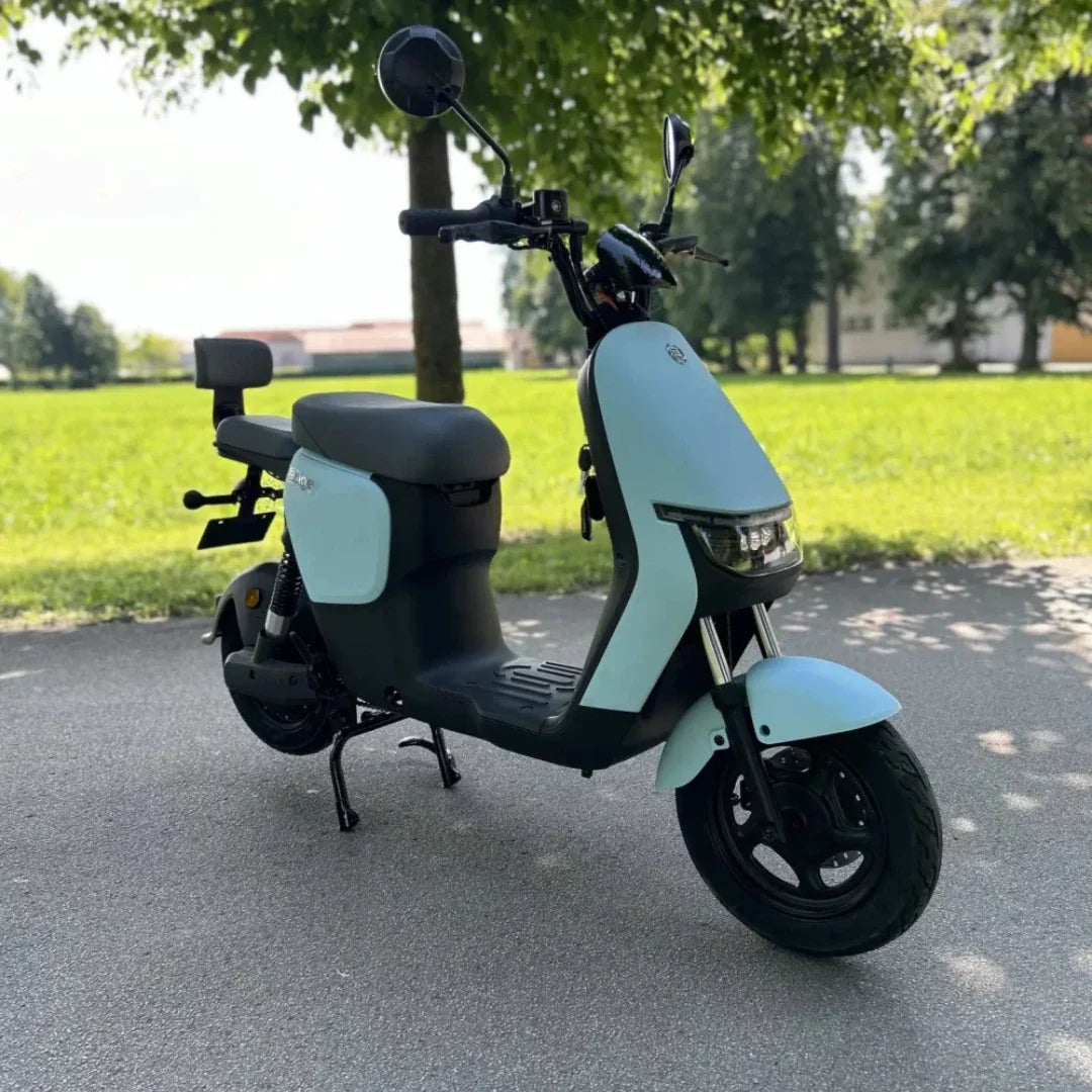 Izdelek na sliki je električni skiro, ki ga odlikuje sodoben in minimalističen dizajn. Znamka in model nista navedena, vendar je skiro opremljen s širokim sedežem in sprednjimi ter zadnjimi ogledali. Na sliki je jasno vidna svetla modra barva karoserije, ki se lepo ujema z zeleno okolico. Skuter je postavljen na asfaltni poti, obdan z drevesi in travo, kar daje občutek svobode in udobja pri vožnji.