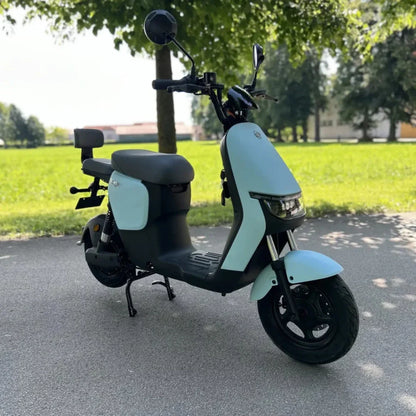 Izdelek na sliki je električni skiro, ki ga odlikuje sodoben in minimalističen dizajn. Znamka in model nista navedena, vendar je skiro opremljen s širokim sedežem in sprednjimi ter zadnjimi ogledali. Na sliki je jasno vidna svetla modra barva karoserije, ki se lepo ujema z zeleno okolico. Skuter je postavljen na asfaltni poti, obdan z drevesi in travo, kar daje občutek svobode in udobja pri vožnji.