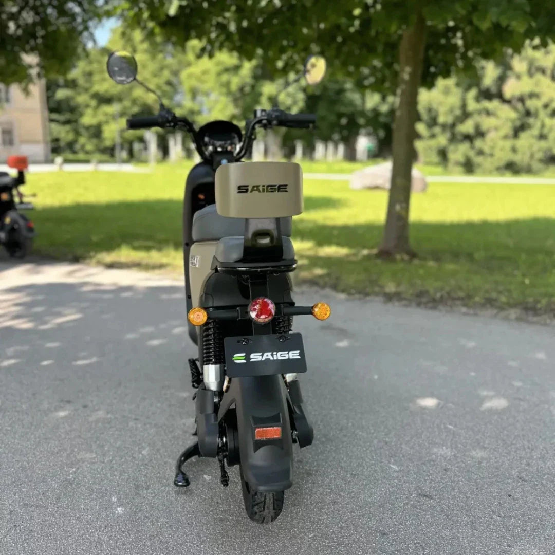 Na sliki je električna motorna kolesa znamke Saige. Vidimo zadnji del motorja, ki ima preprosto obliko in je opremljen z LED lučmi ter oznako znamke. Okolica je zelena, kar nakazuje, da je motor parkiran v naravi, verjetno v parku. Kolo ima tudi klasične vzmetne amortizerje in robustne pnevmatike, kar kaže na njegovo primernost za mestno vožnjo.