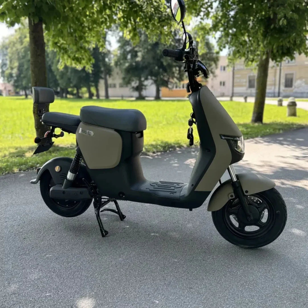 Izdelek na sliki je električni skiro, znamke Stromko. Na sliki je vidna moderna zasnova skiroja, ki ima udobno sedežno površino in široke pnevmatike. Barvna kombinacija je pretežno temno siva z nekaterimi svetlejšimi odtenki. Ozadje prikazuje zeleno okolico, kar nakazuje, da je skiro namenjen vožnji na prostem.