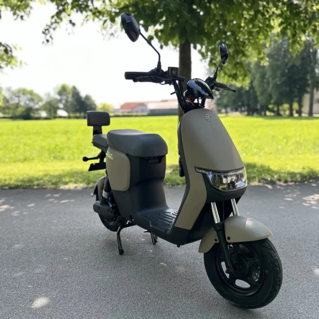 Na sliki je električni skuter, model "Stromko". Ima minimalistično obliko z udobnim sedežem in širokimi ročaji. Barvna kombinacija je v odtenkih sivo-zelene in črne. V ozadju je vidna zelena travnata površina, kar nakazuje na uporabo v naravi ali mestnem okolju. Skuter je zasnovan za enostavno vožnjo in je primeren za mestne poti.