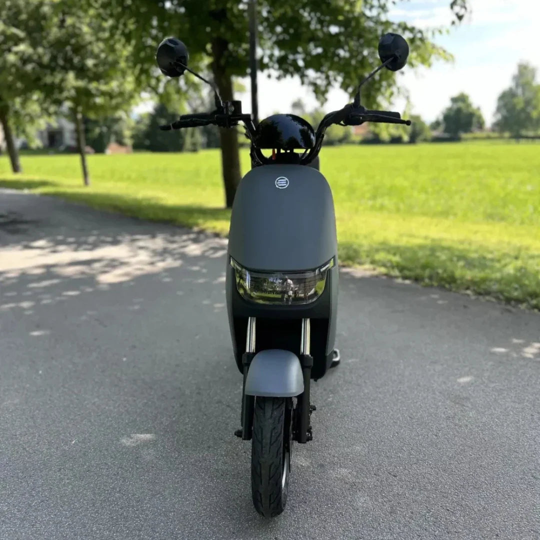 Na sliki je električna motorna kolesa znamke Stromko. Izdelek ima minimalističen in sodoben dizajn, s temno sivo barvo ter sprednjim svetilom. Kolo je postavljeno na asfaltni poti, obdanem z zelenjem, kar daje občutek prostornosti in narave. Na krmilu so vidni tudi ogledali, kar kaže na funkcionalnost in pripravljenost za vožnjo.