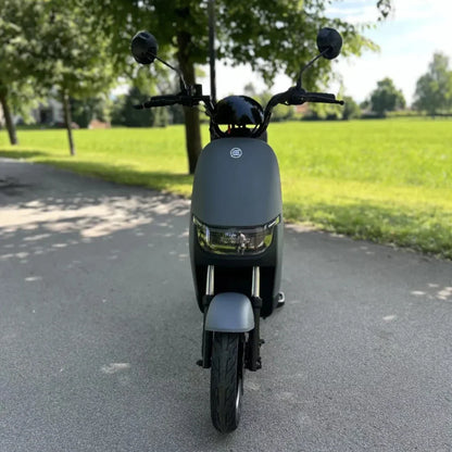 Na sliki je električna motorna kolesa znamke Stromko. Izdelek ima minimalističen in sodoben dizajn, s temno sivo barvo ter sprednjim svetilom. Kolo je postavljeno na asfaltni poti, obdanem z zelenjem, kar daje občutek prostornosti in narave. Na krmilu so vidni tudi ogledali, kar kaže na funkcionalnost in pripravljenost za vožnjo.