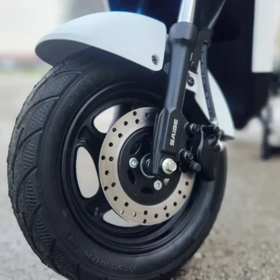 Na sliki je vidna sprednja kolesa električnega motocikla, modela "Stromko". Kolo ima široko pnevmatiko z izrazitim profilom, kar omogoča boljšo oprijemljivost. Vidna je tudi zavorna ploščica in del vzmetenja, kar kaže na robustno zasnovo motocikla. Znamka "SAIGE" je označena na delu zavornega sistema, kar dodatno potrjuje kakovost izdelka.