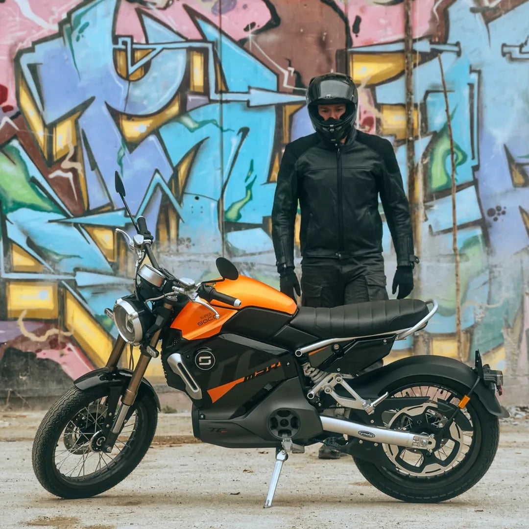 Voznik v črni opremi stoji ob električnem skuterju Super Soco Tc Max ob grafitnem zidu