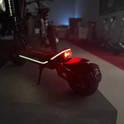 Izdelek na sliki je električni skiro, model Teverun Fighter Mini Pro iz leta 2025. Na sliki je vidna zadnja stran skiroja, ki ima rdečo luč in odsevnike, kar povečuje varnost pri vožnji v temi. Ozadje je nekoliko zamegljeno, kar nakazuje na notranji prostor, kjer so vidni tudi drugi skiroji.