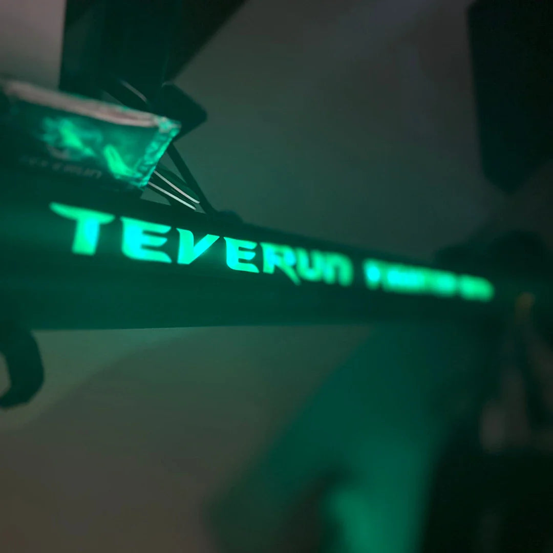 Izdelek na sliki je električni skiro, model Teverun Fighter Mini Pro iz leta 2025. Na sliki je vidna osvetljena oznaka znamke Teverun na okvirju skiroja, ki daje sodoben in atraktiven videz. V ozadju je tudi del skiroja, kar nakazuje na njegovo zasnovo in funkcionalnost.