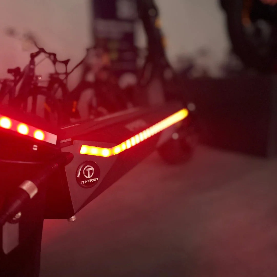 Na sliki je električni skiro znamke Teverun, model Fighter Mini Pro 2025. Vidna je zadnja stran skiroja, kjer so nameščene LED luči, ki osvetljujejo pot. Skuter ima robustno konstrukcijo in sodoben dizajn, kar nakazuje na njegovo funkcionalnost in zmogljivost.