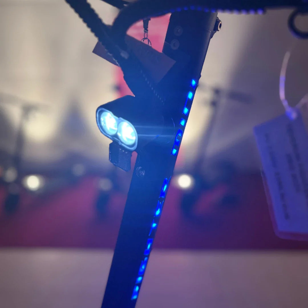 Izdelek na sliki je električni skiro, model Teverun Fighter Mini Pro iz leta 2025. Na sliki je vidna osvetlitev na krmilu skiroja, ki vključuje dve svetli LED luči ter modre LED trakove, kar daje skiroju sodoben in atraktiven videz. V ozadju so vidni tudi drugi skiroji, kar nakazuje, da gre za razstavo ali prodajno mesto.