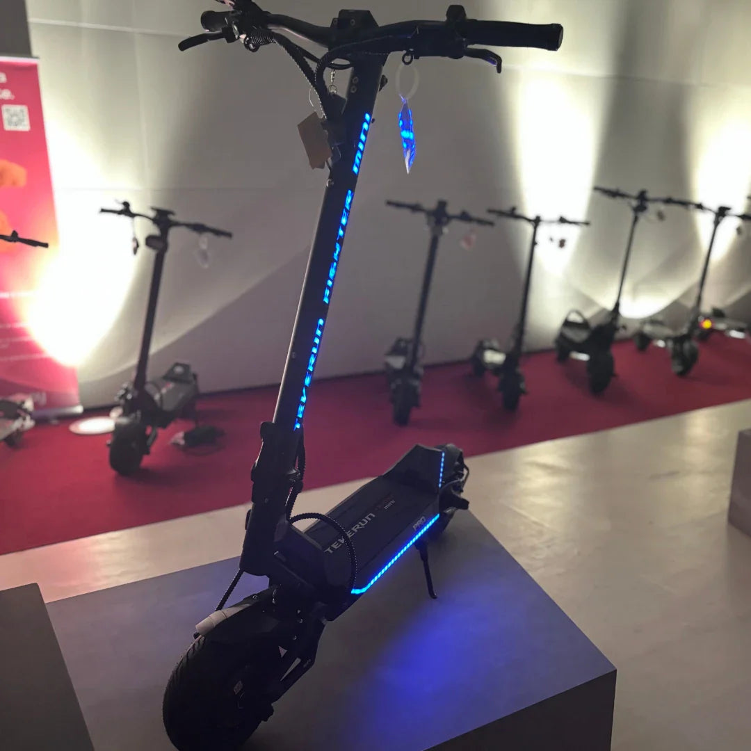 Na sliki je električni skiro znamke Teverun, model Fighter Mini Pro 2025. Izdelek je predstavljen na stojalu, z vidnimi LED lučmi, ki dodajajo estetski videz. V ozadju so še drugi skiroji iste znamke, kar nakazuje na razstavo ali predstavitev.