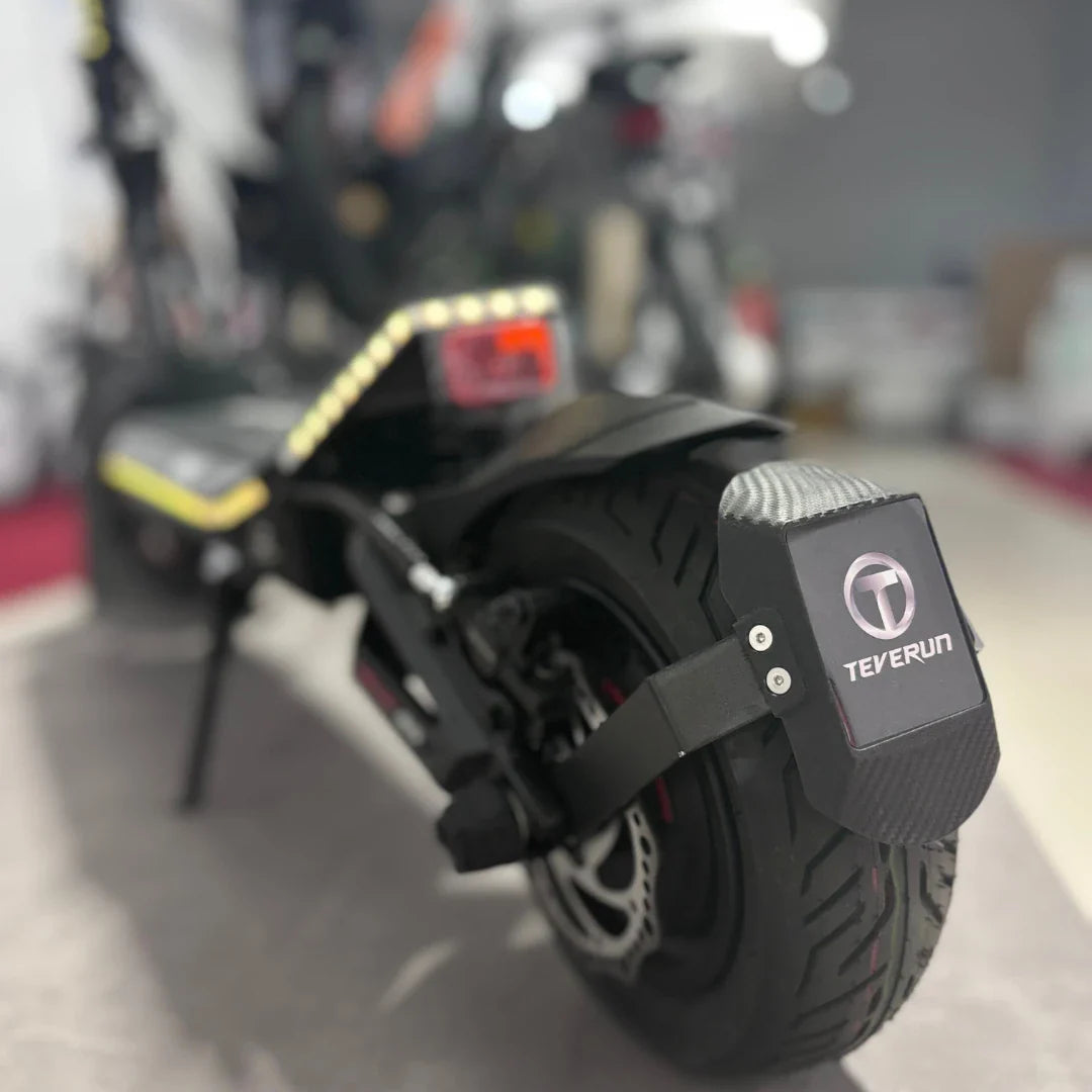 Na sliki je električni skiro znamke Teverun, model Fighter Mini Pro 2025. Vidna je zadnja stran skiroja, kjer se nahaja široko kolo z značilnim profilom in zaščitno ploščico, na kateri je logotip Teverun. Opremljen je tudi z LED lučmi, kar povečuje varnost pri vožnji v temi.