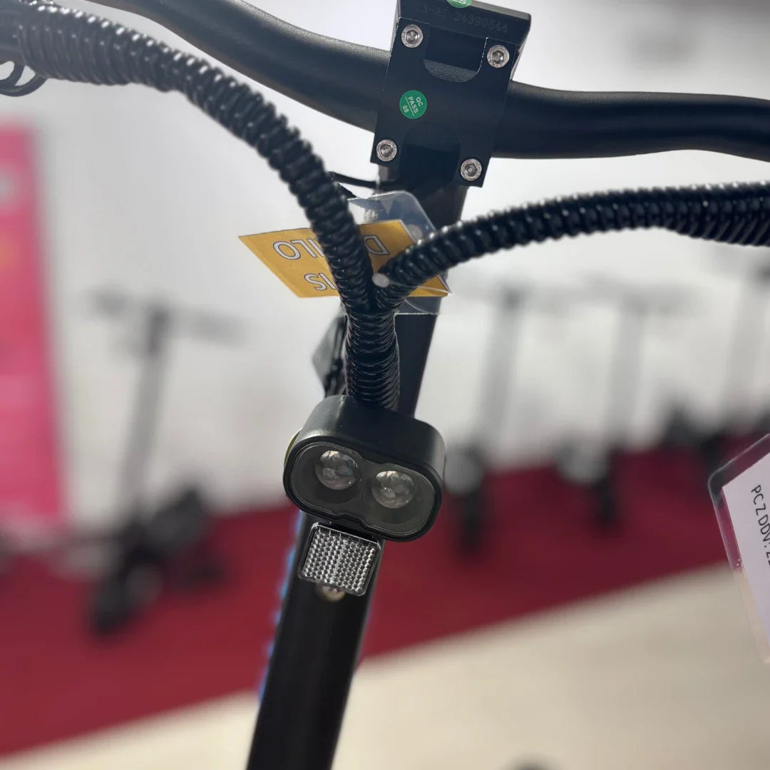 Na sliki je električni skiro, model Teverun Fighter Mini Pro 2025. Vidna je sprednja stran skiroja, kjer so nameščeni ročaji in luči. Luči so zasnovane za osvetlitev ceste, kar povečuje varnost pri vožnji v temi. V ozadju so vidni tudi drugi skiroji, kar nakazuje, da gre za prodajno mesto.