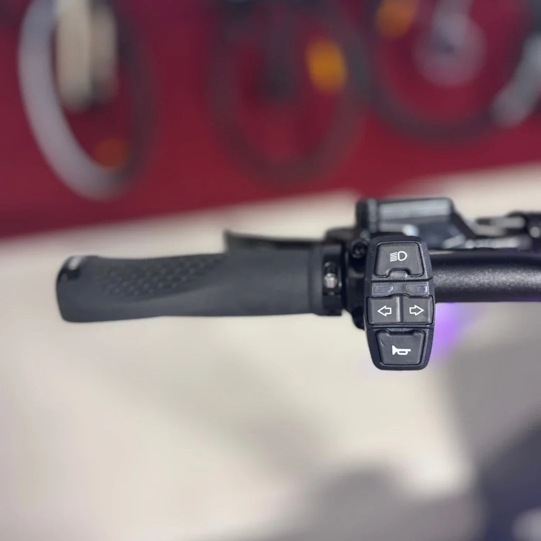 Na sliki je prikazan električni skiro, model Teverun Fighter Mini Pro 2025. Vidni so del krmila z ročajem in kontrolnimi gumbi, ki vključujejo funkcije za luči in smerokaze. Ozadje je nejasno, vendar nakazuje na okolje, kjer so morda razstavljeni tudi drugi podobni izdelki.