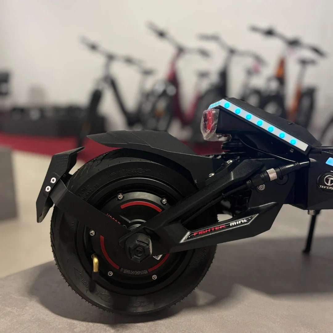 Izdelek na sliki je električni skiro znamke Teverun, model Fighter Mini Pro 2025. Na sliki je vidna zadnja stran skiroja, kjer se lahko opazi robustna konstrukcija, široka pnevmatika ter LED luči, ki so nameščene na zadnjem delu. V ozadju so vidni tudi drugi električni skiroji, kar nakazuje, da gre za prodajno mesto ali razstavo.