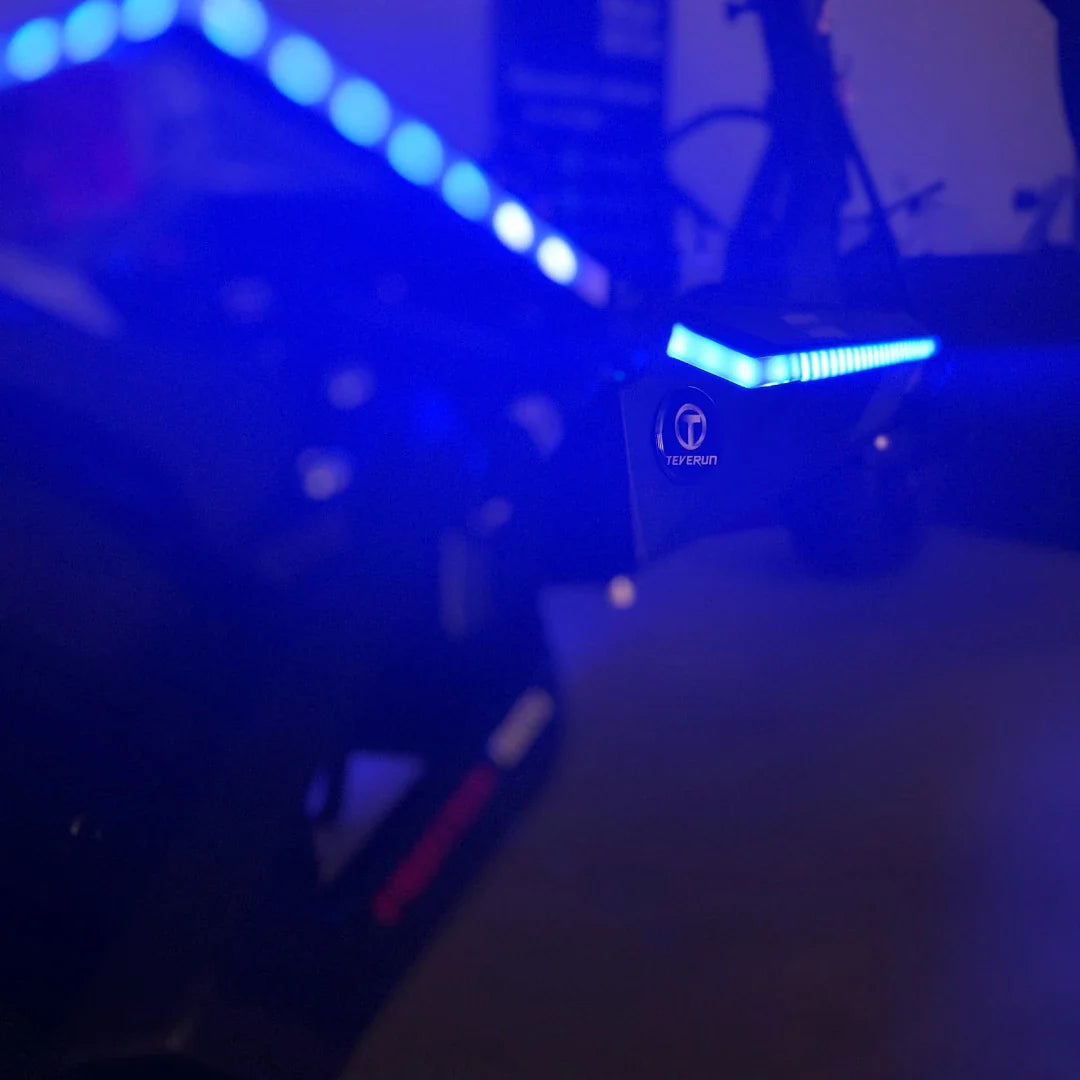 Na sliki je električni skiro, model Teverun Fighter Mini Pro iz leta 2025. Vidna je del skiroja s svetlobnimi LED trakovi, ki oddajajo modro svetlobo, ter logotip znamke Teverun. Ozadje je nekoliko zamegljeno, kar poudari detajle skiroja.
