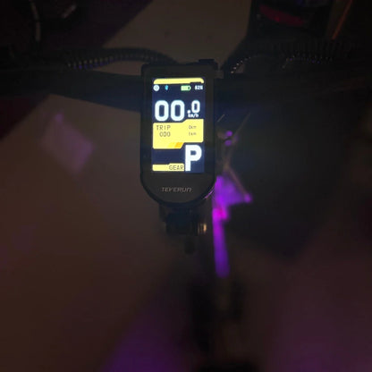 Na sliki je električni skiro, model Teverun Fighter Mini Pro 2025. Vidna je kontrolna enota na krmilu, ki prikazuje hitrost, skupno prevoženo razdaljo in stanje baterije. Zaslon ima tudi oznako za prestavo, kar nakazuje na možnost prilagajanja hitrosti. Osvetlitev v ozadju dodaja sodoben videz.