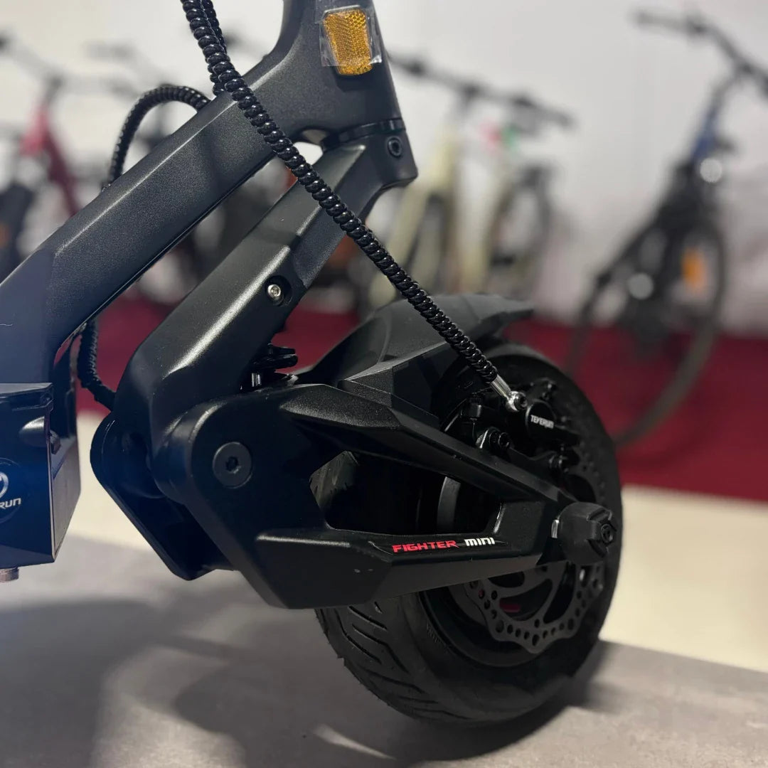 Izdelek na sliki je električni skiro, znamke Teverun, model Fighter Mini Pro 2025. Na sliki je prikazan del zadnjega dela skiroja, vključno z motorjem in kolesom. Vidna je robustna konstrukcija, črne barve, ter detajli, kot so zavorni sistem in zaščitne komponente. V ozadju so vidni tudi drugi kolesarski izdelki.