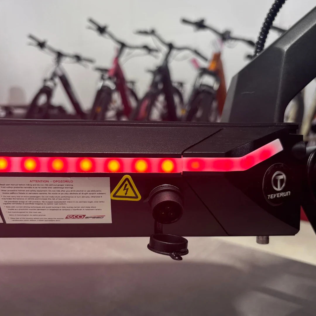 Izdelek na sliki je električni skiro, model Teverun Fighter Mini Pro iz leta 2025. Na sliki je prikazana zadnja stran skiroja, kjer so vidne rdeče LED luči, priključek za polnjenje ter opozorilna oznaka za električno energijo. V ozadju so vidni tudi drugi električni kolesi.