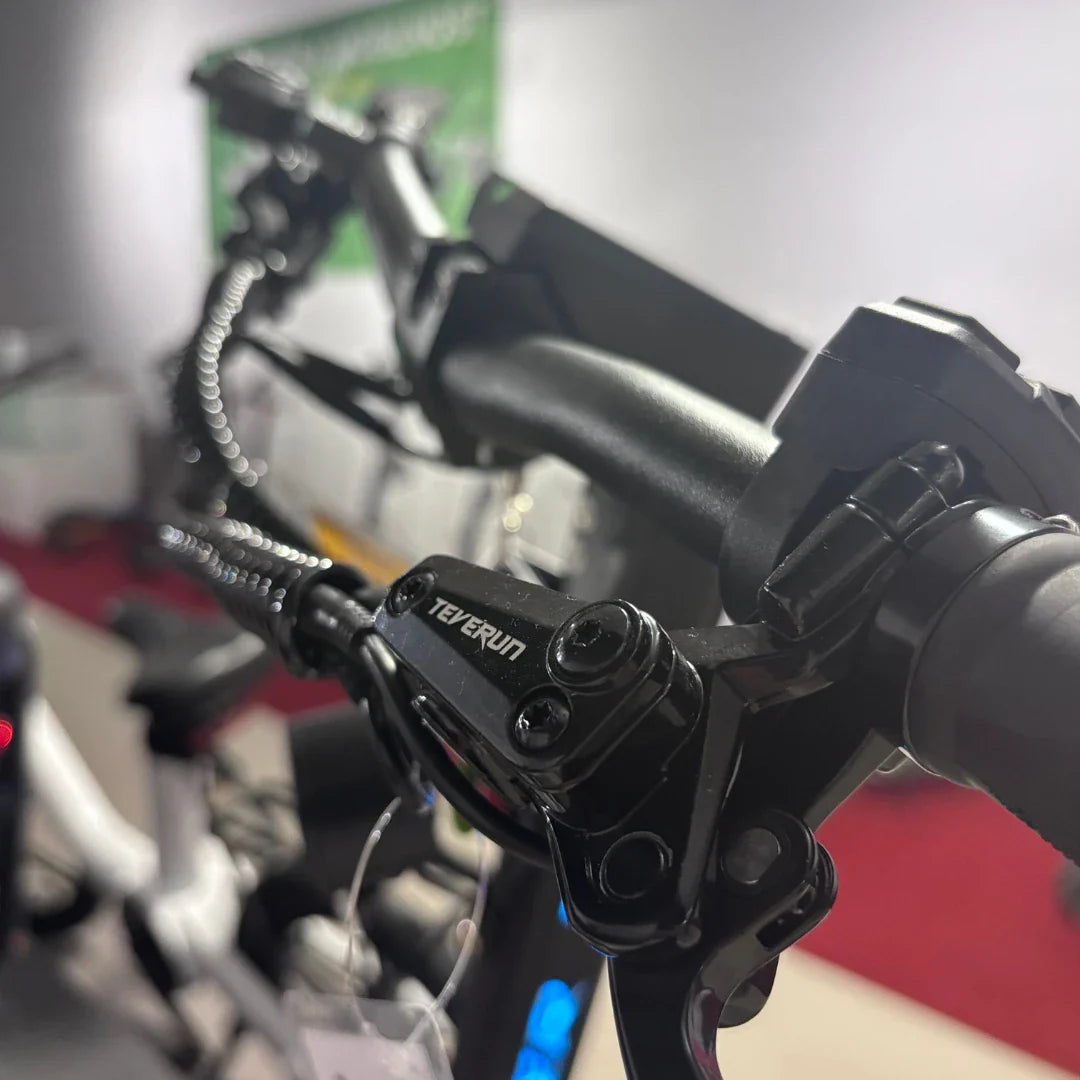 Izdelek je električni skiro, model Teverun Fighter Mini Pro 2025. Na sliki je vidna ročica za zaviranje, ki je del krmila skiroja. Oprema je moderna in robustna, kar kaže na kakovost izdelka. V ozadju je del prostora, kjer je skiro razstavljen.