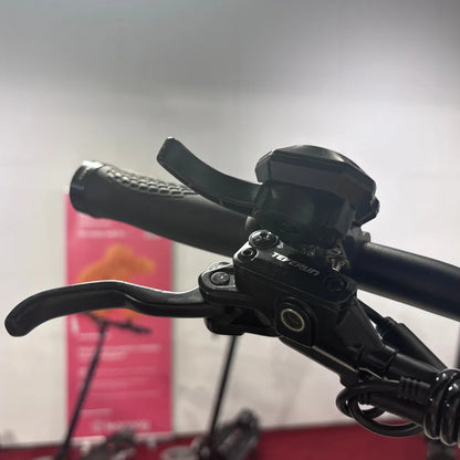 Izdelek je električni skiro, model Teverun Fighter Mini Pro 2025. Na sliki je vidna ročica za zaviranje in del krmila, ki sta narejena iz črnega materiala. Ročica je ergonomsko oblikovana, kar omogoča udobno uporabo. V ozadju so delčki drugega skiroja, kar nakazuje, da je slika posneta v prodajnem prostoru ali razstavišču.