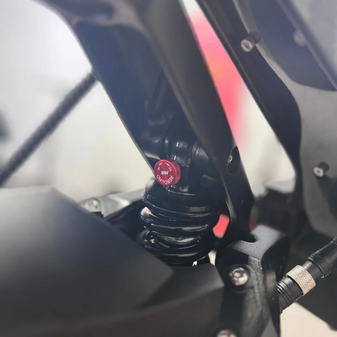 Izdelek na sliki je električni skiro, model Teverun Fighter Mini Pro 2025. Na sliki je vidna podrobnost vzmetenja, kjer je prikazana črna vzmet s rdečim nastavitvenim gumbom. Ta gumb omogoča prilagoditev trdote vzmetenja, kar prispeva k udobni vožnji.