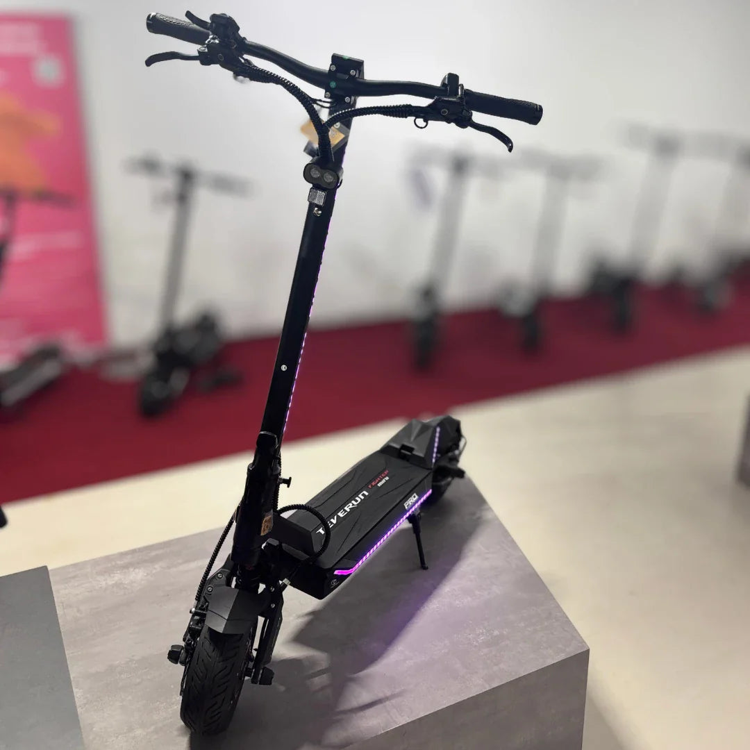 Izdelek na sliki je električni skiro, model Teverun Fighter Mini Pro 2025. Na sliki je vidna sprednja stran skiroja, ki ima robustno konstrukcijo in športen dizajn. Opremljen je z dvema ročajema za upravljanje ter LED lučmi, kar daje sodoben videz. V ozadju so vidni tudi drugi električni skiroji, kar nakazuje, da gre za razstavni prostor.