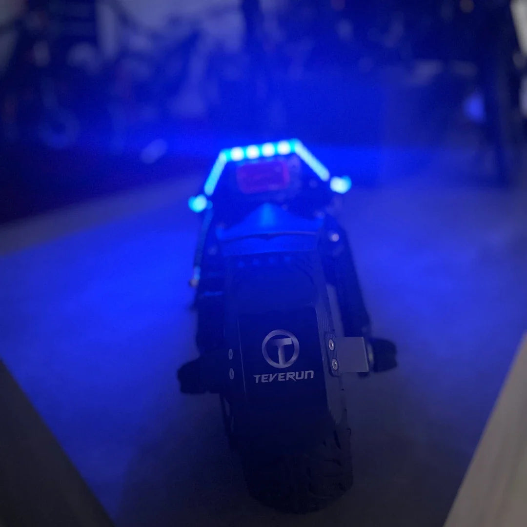 Izdelek na sliki je električni skiro, model Teverun Fighter Mini Pro 2025. Na sliki je vidna zadnja stran skiroja, ki ima svetleče modre LED luči ter značko znamke Teverun. Skuter se zdi robusten, s širokimi pnevmatikami, kar nakazuje na dobro stabilnost in zmogljivost.