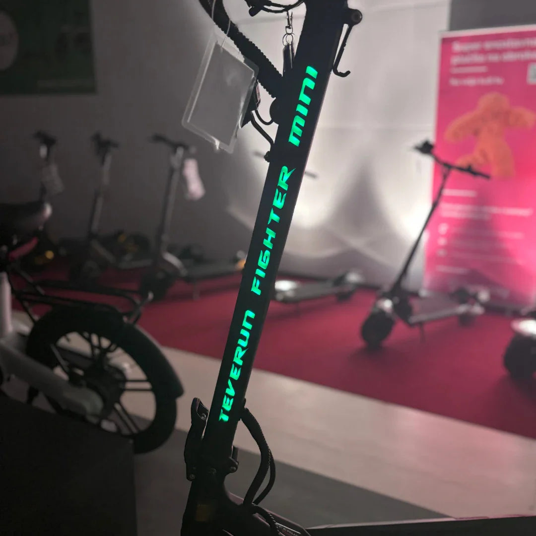 Izdelek na sliki je električni skiro, model Teverun Fighter Mini iz leta 2025. Na sliki je vidna vertikalna cev skiroja z imenom modela, ki je osvetljena v zeleni barvi. V ozadju so vidni tudi drugi električni skiroji, kar nakazuje na razstavo ali prodajno mesto.