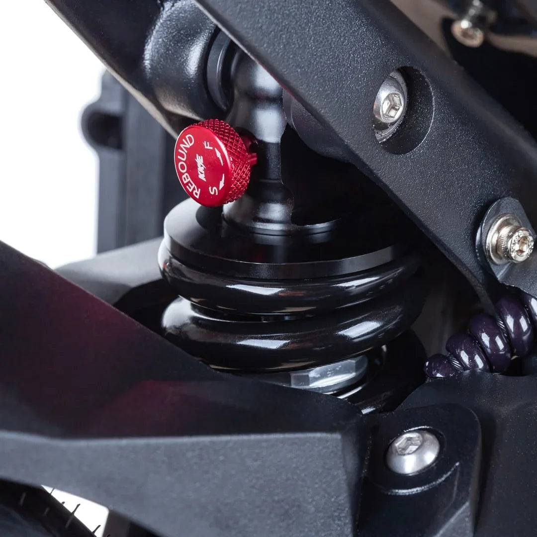 Izdelek je električni skiro, model Teverun Fighter Mini Pro 2025. Na sliki je prikazana podrobnost vzmetenja, ki vključuje nastavljiv sistem za povratno delovanje, obdan s črno in rdečo barvo. Vidni so tudi deli okvirja in mehanizma, kar kaže na robustno zasnovo skiroja.