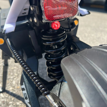 Na sliki je električni skiro, model Teverun Fighter Mini Pro 2025. Vidna je zadnja vzmet, ki je črna in omogoča boljšo absorpcijo udarcev, ter rdeča odsevna lučka, ki povečuje varnost pri vožnji. Oprema skiroja vključuje tudi zaščitni kabel, kar kaže na premišljeno zasnovo za trajnost in varnost.