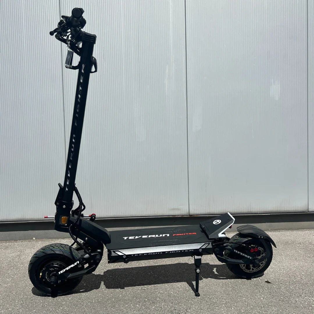 Na sliki je električni skiro, model Teverun Fighter Mini Pro 2025. Izdelek ima robustno konstrukcijo, široko platformo za stojišče in velike pnevmatike, kar nakazuje na dobro stabilnost in udobje med vožnjo. Skuter je črne barve z belimi detajli in logotipom znamke, kar mu daje sodoben in športen videz.