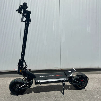 Na sliki je električni skiro, model Teverun Fighter Mini Pro 2025. Izdelek ima robustno konstrukcijo, široko platformo za stojišče in velike pnevmatike, kar nakazuje na dobro stabilnost in udobje med vožnjo. Skuter je črne barve z belimi detajli in logotipom znamke, kar mu daje sodoben in športen videz.