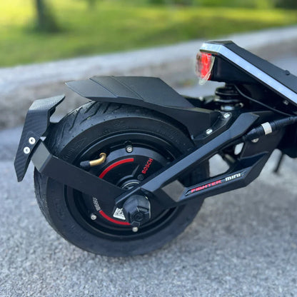 Na sliki je električni skiro znamke Teverun, model Fighter Mini Pro 2025. Vidna je zadnja kolesa skiroja, kjer se nahajajo elementi, kot so motor, zavora in zaščitni del. Kolo je opremljeno z robustnim gumijastim plaščem, kar omogoča boljšo oprijemljivost na različnih površinah. Skupna zasnova nakazuje na sodobno in športno naravnanost izdelka.