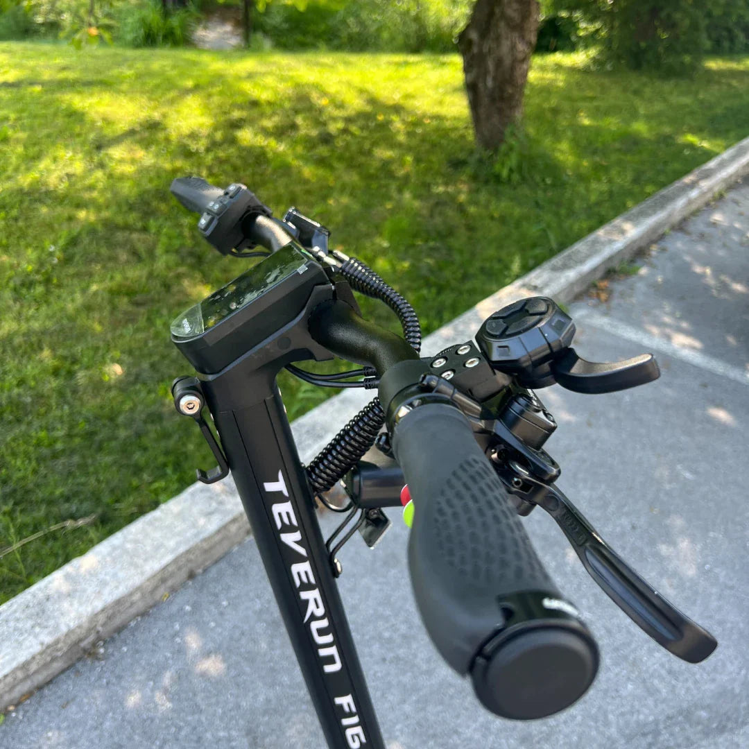 Izdelek na sliki je električni skiro, model Teverun Fighter Mini Pro iz leta 2025. Na sliki so vidni ročaji skiroja z nadzorno ploščo, ki prikazuje različne funkcije, ter gumbi za upravljanje. Opremljen je z ergonomskimi ročaji in dodatnimi kontrolami, kar nakazuje na poudarek na udobju in funkcionalnosti med vožnjo. V ozadju je zeleno okolje, kar daje občutek prostornosti in narave.