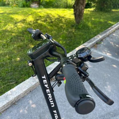 Izdelek na sliki je električni skiro, model Teverun Fighter Mini Pro iz leta 2025. Na sliki so vidni ročaji skiroja z nadzorno ploščo, ki prikazuje različne funkcije, ter gumbi za upravljanje. Opremljen je z ergonomskimi ročaji in dodatnimi kontrolami, kar nakazuje na poudarek na udobju in funkcionalnosti med vožnjo. V ozadju je zeleno okolje, kar daje občutek prostornosti in narave.