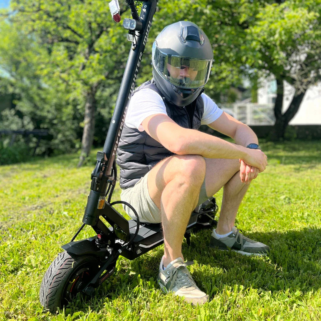 Na sliki je električni skiro znamke Teverun, model Fighter Mini Pro 2025. Izdelek je prikazan na travnati površini, ob njem sedi oseba v čeladi in brezrokavniku. Skuter ima robustno zasnovo, široke pnevmatike ter je opremljen s prednjim in zadnjim svetilom. V ozadju so vidne drevesne krošnje in del stavbe, kar nakazuje na zunanje okolje.