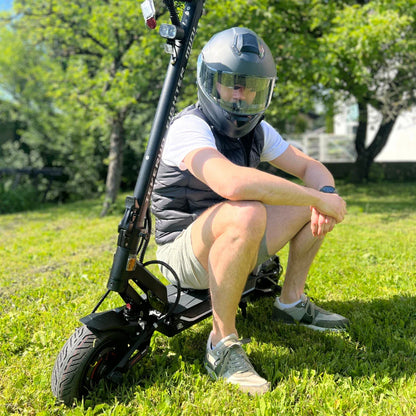 Na sliki je električni skiro znamke Teverun, model Fighter Mini Pro 2025. Izdelek je prikazan na travnati površini, ob njem sedi oseba v čeladi in brezrokavniku. Skuter ima robustno zasnovo, široke pnevmatike ter je opremljen s prednjim in zadnjim svetilom. V ozadju so vidne drevesne krošnje in del stavbe, kar nakazuje na zunanje okolje.