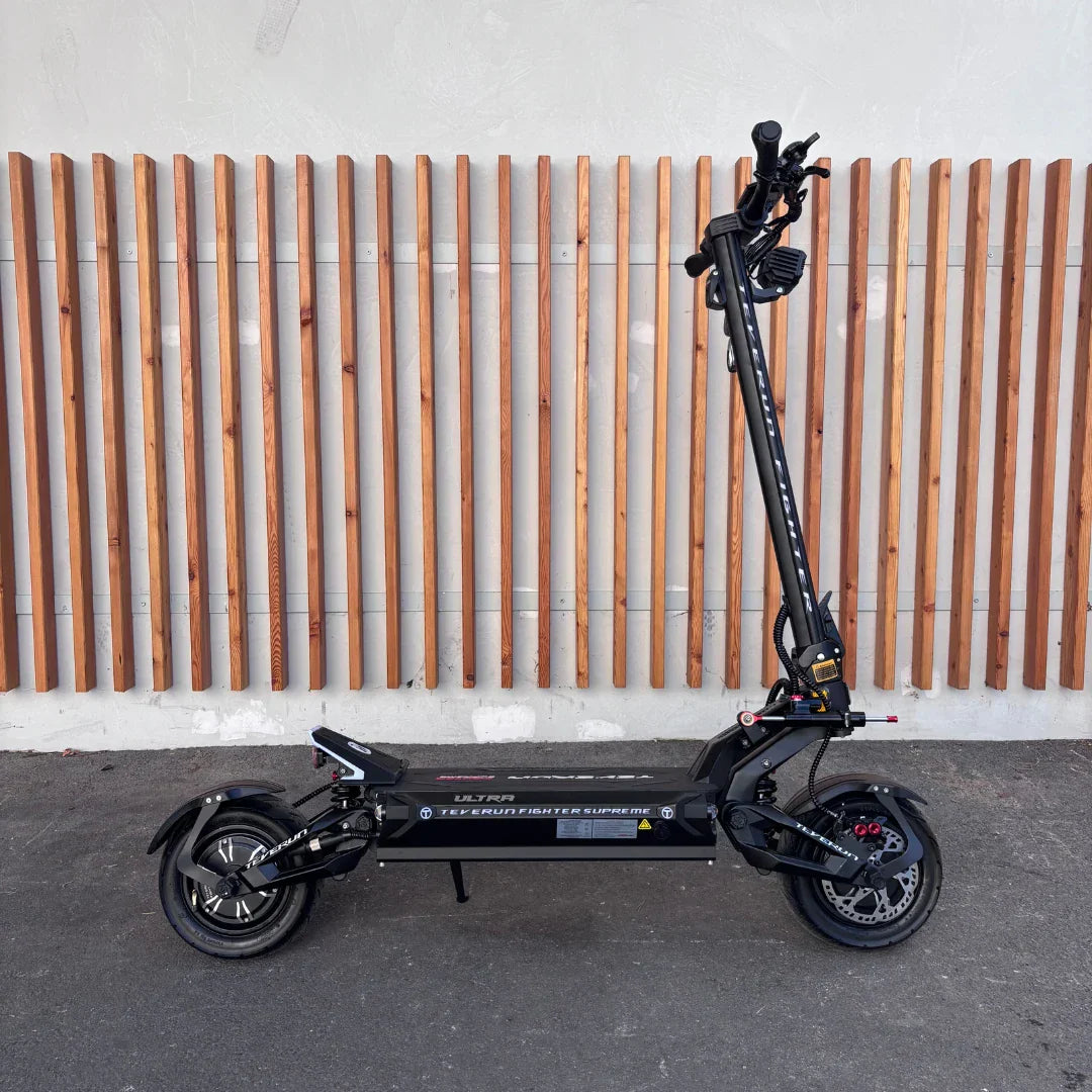 Izdelek na sliki je električni skiro, znamke Teverun, model Fighter Supreme 7260 Ultra. Na sliki je skiro postavljen na trdno podlago, s črnimi okvirji in širokimi kolesi, kar nakazuje na robustno zasnovo. Vidna je tudi krmilo z različnimi kontrolami in zavorami, kar nakazuje na njegovo zmogljivost in funkcionalnost. V ozadju je lesena stena, ki daje sliki estetski pridih.