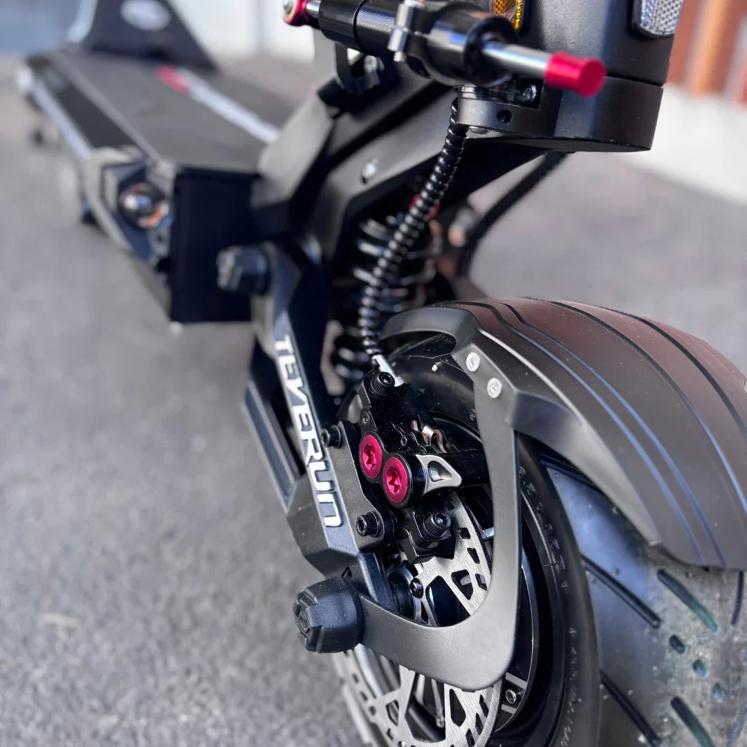 Izdelek na sliki je električni skiro, model Teverun Fighter Supreme 7260 Ultra. Na sliki je prikazan del skiroja, ki vključuje robustno zadnje kolo s široko pnevmatiko ter disk zavora. Vidni so tudi detajli, kot so vijaki v rdeči barvi in vzmetenje, kar nakazuje na visoko kakovost in zmogljivost skiroja.