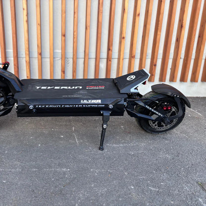 Izdelek je električni skiro, model Teverun Fighter Supreme 7260 Ultra. Na sliki je prikazan skiro v črni barvi z značilnim logotipom znamke Teverun. Ima široko platformo, velike kolesa in stabilno zasnovo, kar nakazuje na robustnost in zmogljivost. Vidna je tudi podpora za parkiranje, kar omogoča enostavno postavitev skiroja.