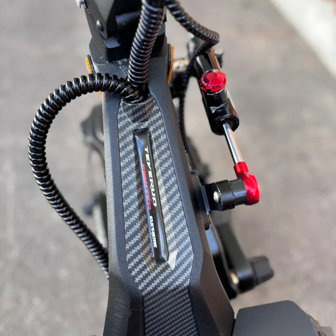 Izdelek na sliki je električni skiro, znamke Teverun, model Fighter Supreme 7260 Ultra. Na sliki je vidna podrobnost skiroja, ki vključuje ogljikovo vlakno na okvirju ter rdeč nastavljiv gumb, ki verjetno služi za prilagajanje nekaterih funkcij. Celoten videz nakazuje na robustno in zmogljivo zasnovo.