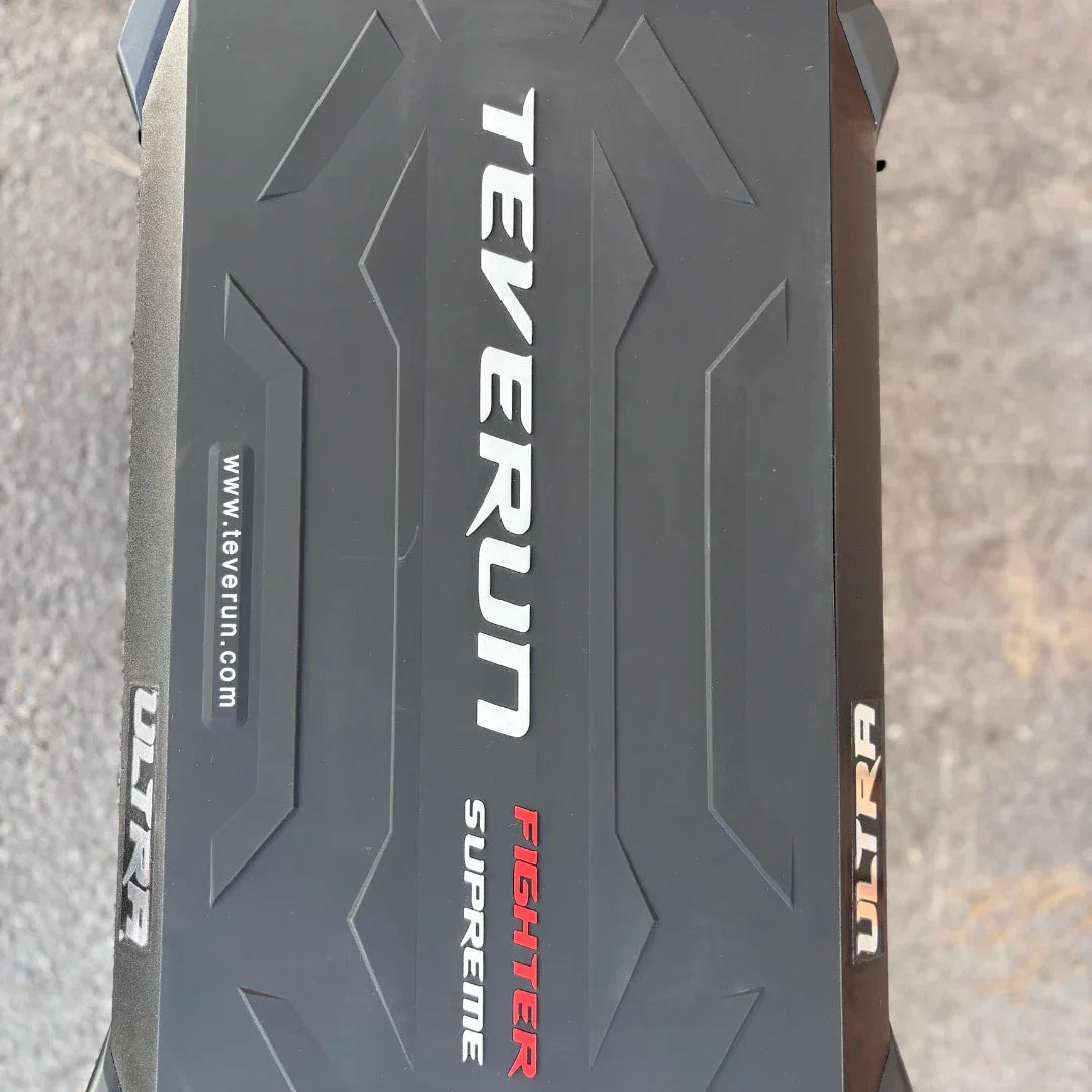 Izdelek je električni skiro, znamke Teverun, model Fighter Supreme ULTRA. Na sliki je vidna zgornja površina skiroja, ki ima črno teksturo z izbočenimi vzorci ter napisom "TEVERUN" in "FIGHTER SUPREME ULTRA" v različnih barvah. Površina je zasnovana za dober oprijem, kar je pomembno za varno vožnjo.