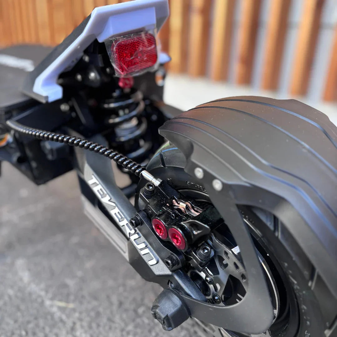 Izdelek je električni skiro, znamke Teverun, model Fighter Supreme 7260 Ultra. Na sliki je prikazan zadnji del skiroja, kjer so vidni amortizer, zadnja luč ter del kolesa z zavorami. Detajli vključujejo tudi črno in rdečo barvno kombinacijo ter robustno zasnovo, kar nakazuje na visoko zmogljivost in stabilnost.