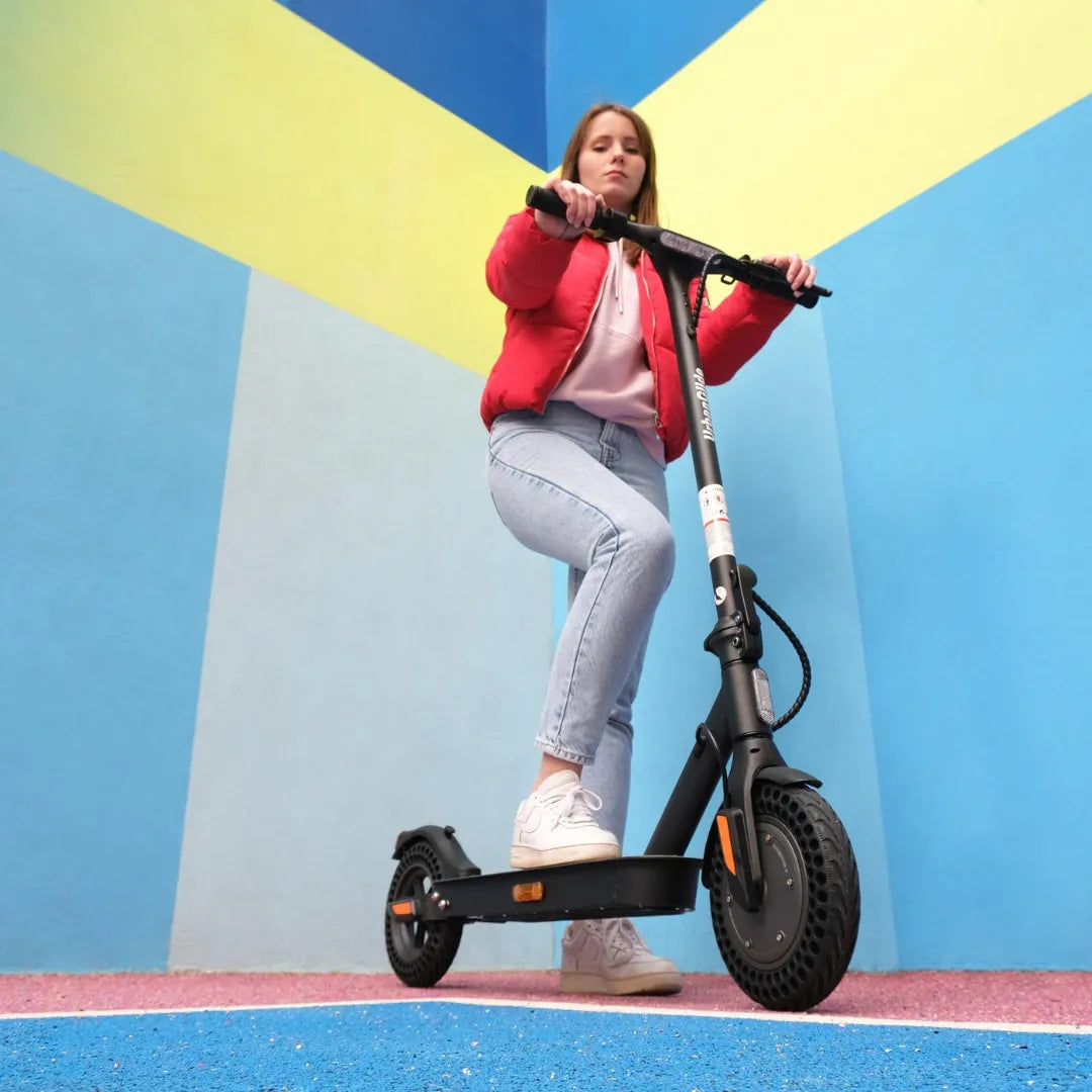 Oseba se vozi z električnim skirojem Urbanride 100xs pred pisano steno.