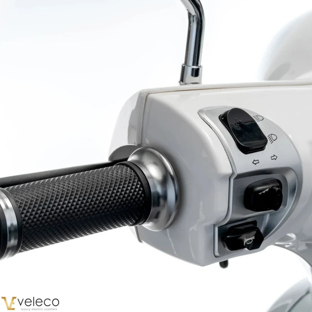 Na sliki je električni skuter, model Veleco Cristal. Vidni so ročaji in del nadzorne plošče, kjer so gumbi za luči in smerokaze. Skuter ima eleganten dizajn z gladko belo površino in teksturirane gumijaste ročaje, kar omogoča udoben oprijem.