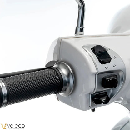Na sliki je električni skuter, model Veleco Cristal. Vidni so ročaji in del nadzorne plošče, kjer so gumbi za luči in smerokaze. Skuter ima eleganten dizajn z gladko belo površino in teksturirane gumijaste ročaje, kar omogoča udoben oprijem.
